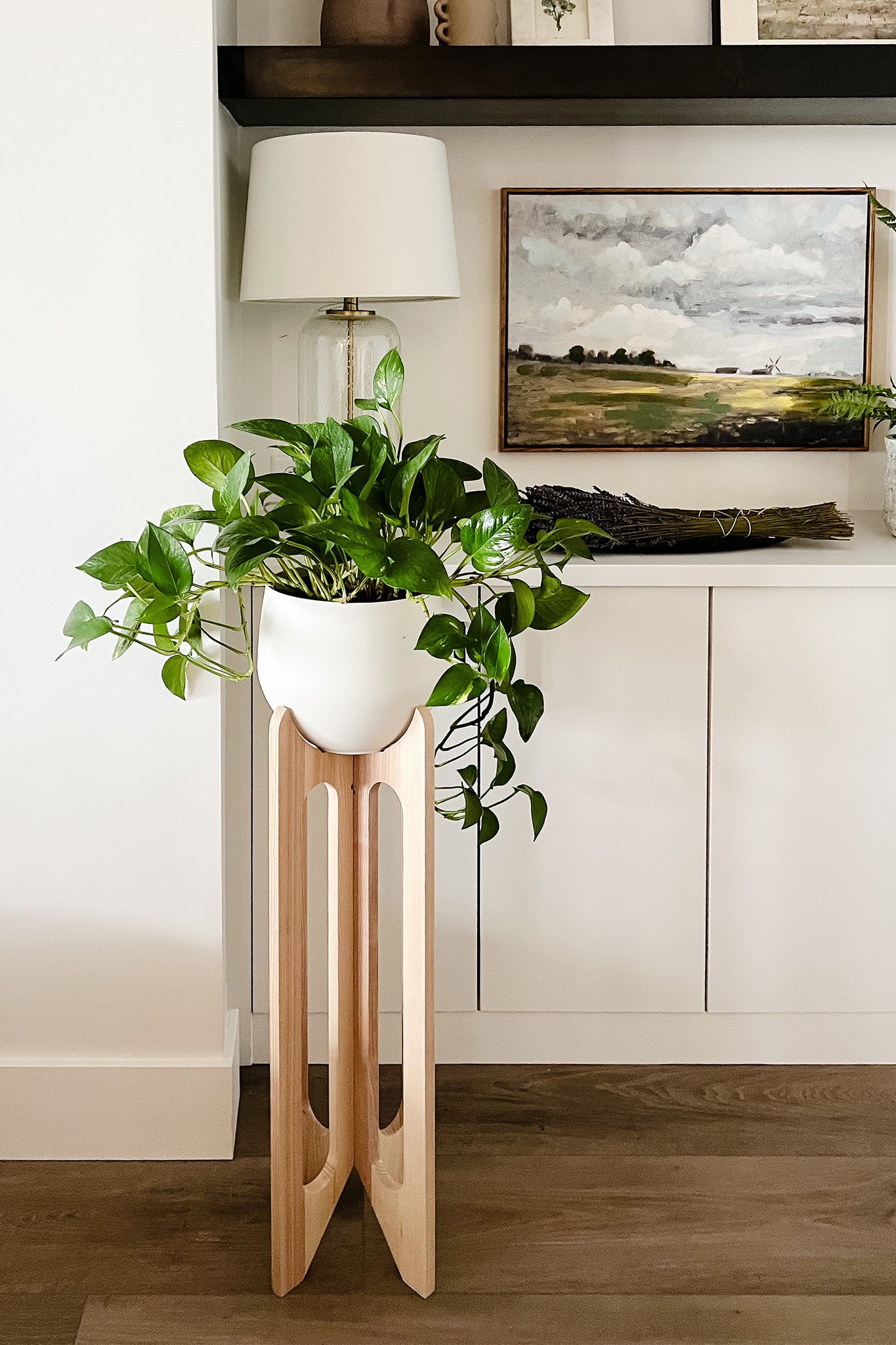 An Arches Plant Stand in a modern living room in front of built in bookshelves with decorative elements like framed artwork and a lamp.