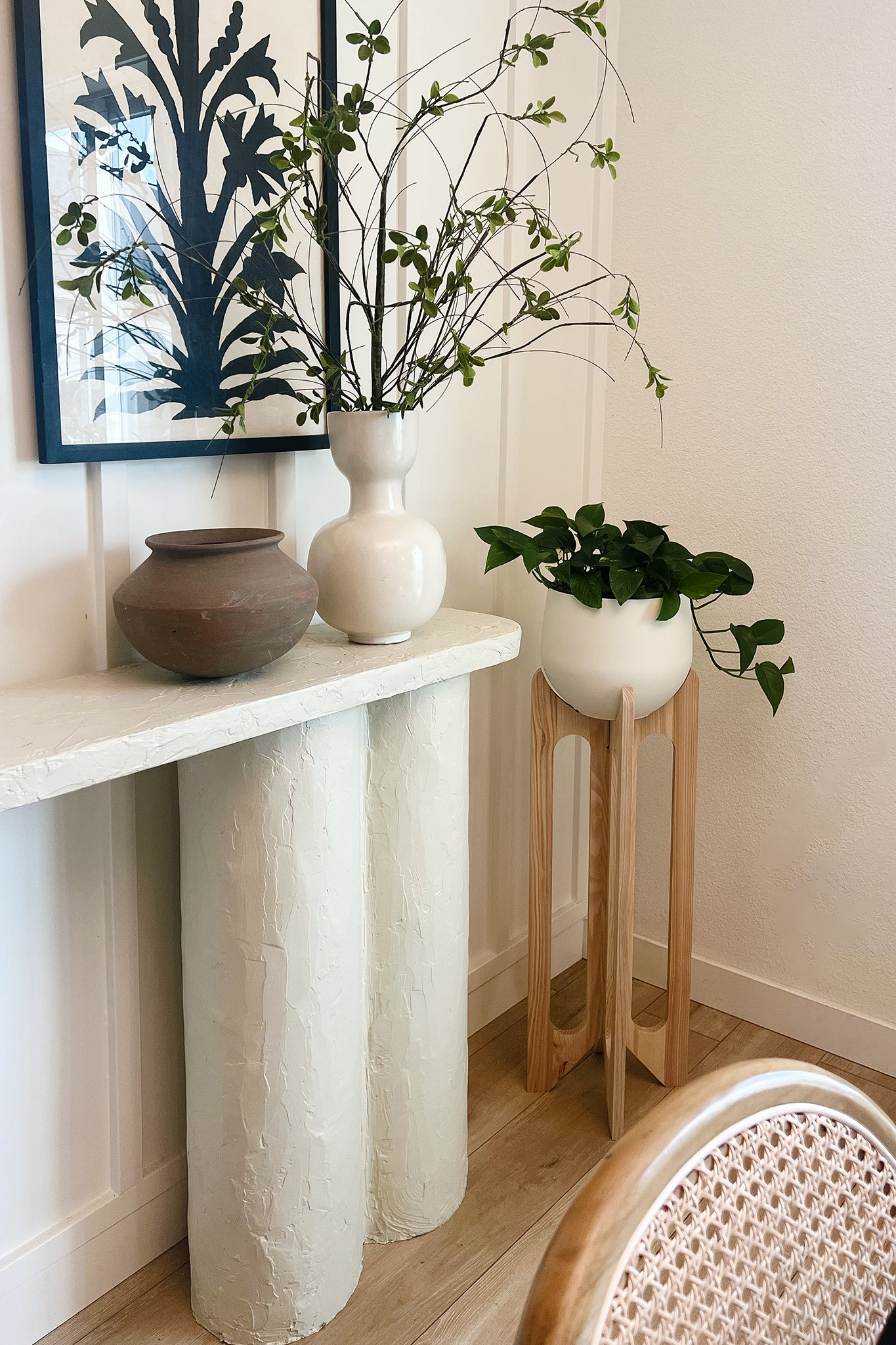 A white console table with vases on pottery on top with the Arches Plant Stand next to it. There is framed artwork on the wall above the console table.