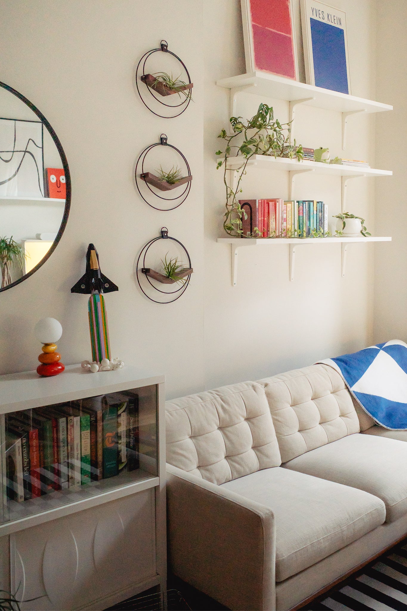 A modern living room with a gray sofa, white walls, colorful books, and three wall-mounted circular air plant hangers in a vertical line on the wall next to a round mirror.