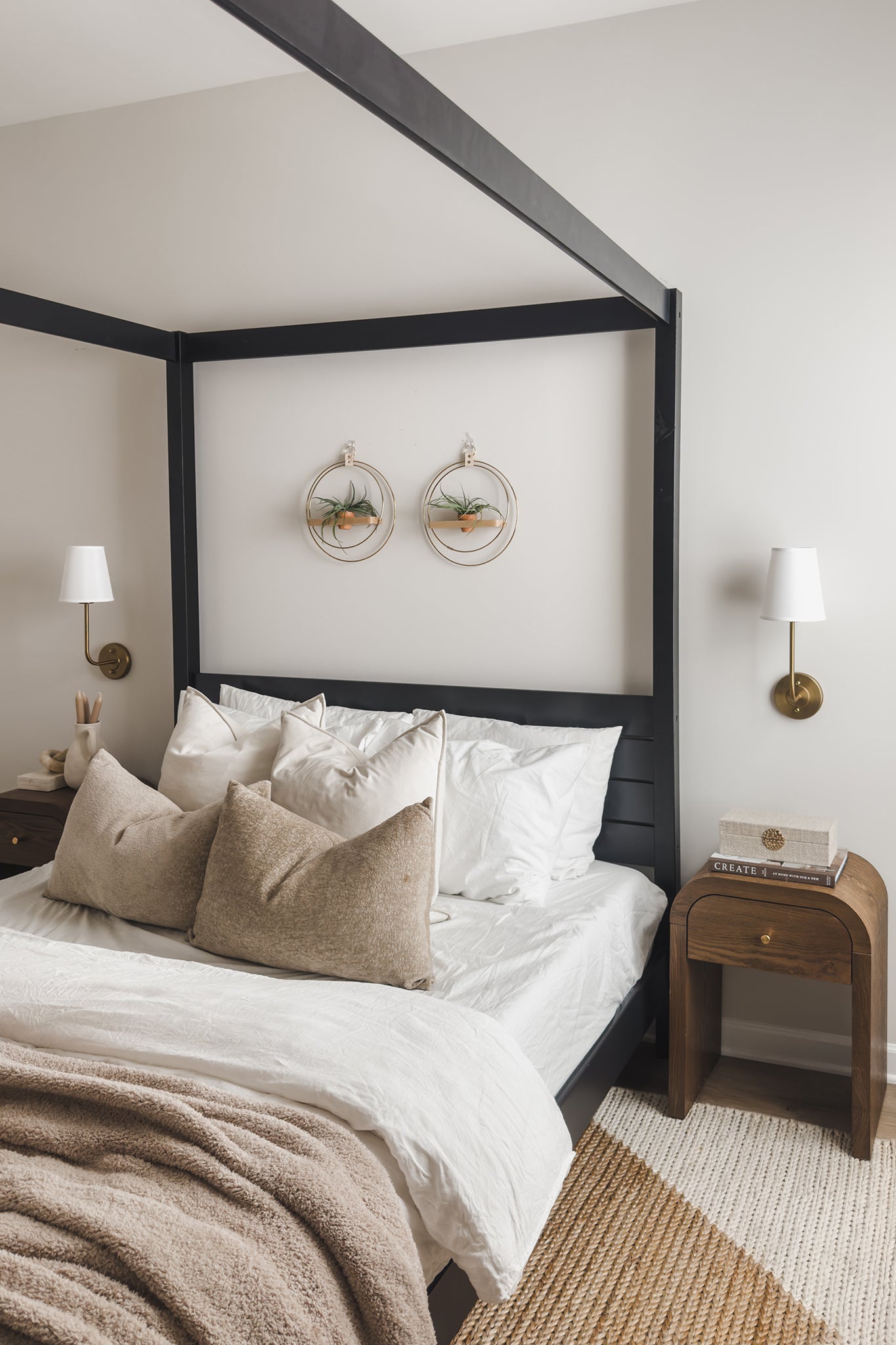 Neatly made bed with white bedding and brown throw blanket in a bedroom setting with two wall-mounted circular planters with air plants in a terra cotta pot on the wall above.