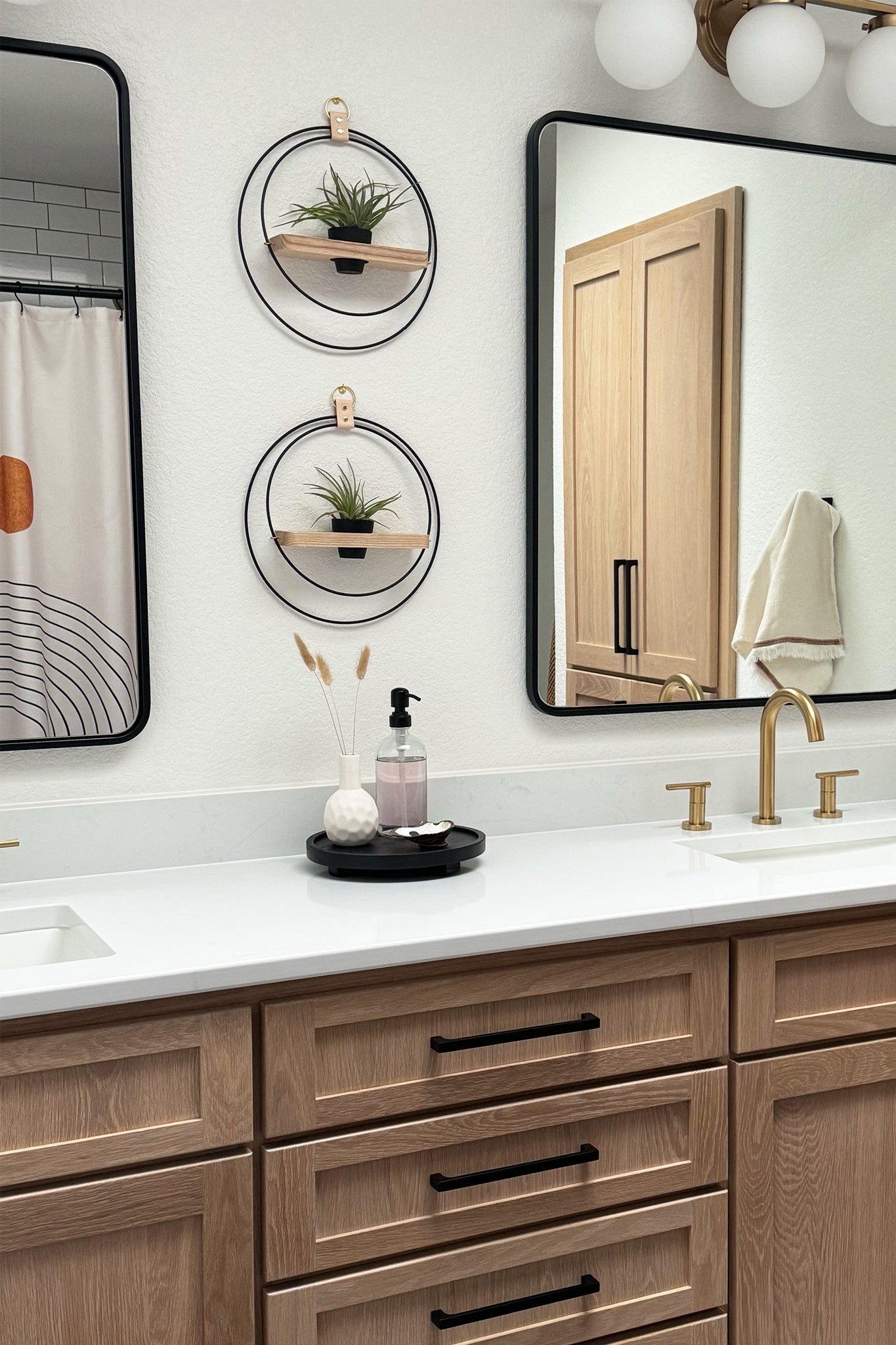 A modern bathroom with white countertops with wood cabinets with two Wall-mounted circular planters placed between the two black rimmed vanity mirrors.