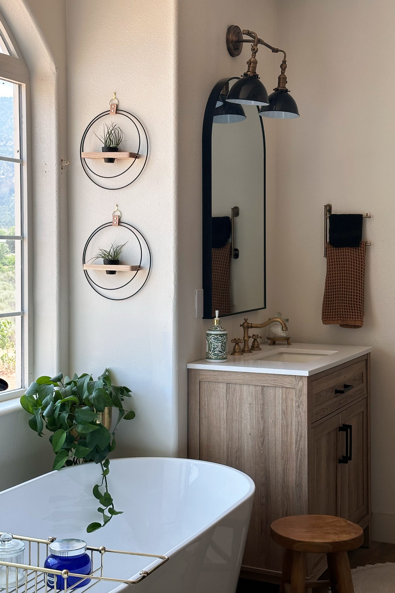 Bathroom interior with a freestanding bathtub, wooden vanity, and two wall-mounted circular planters with air plants in a terra cotta pot a neutral wall next to a large, arched window.