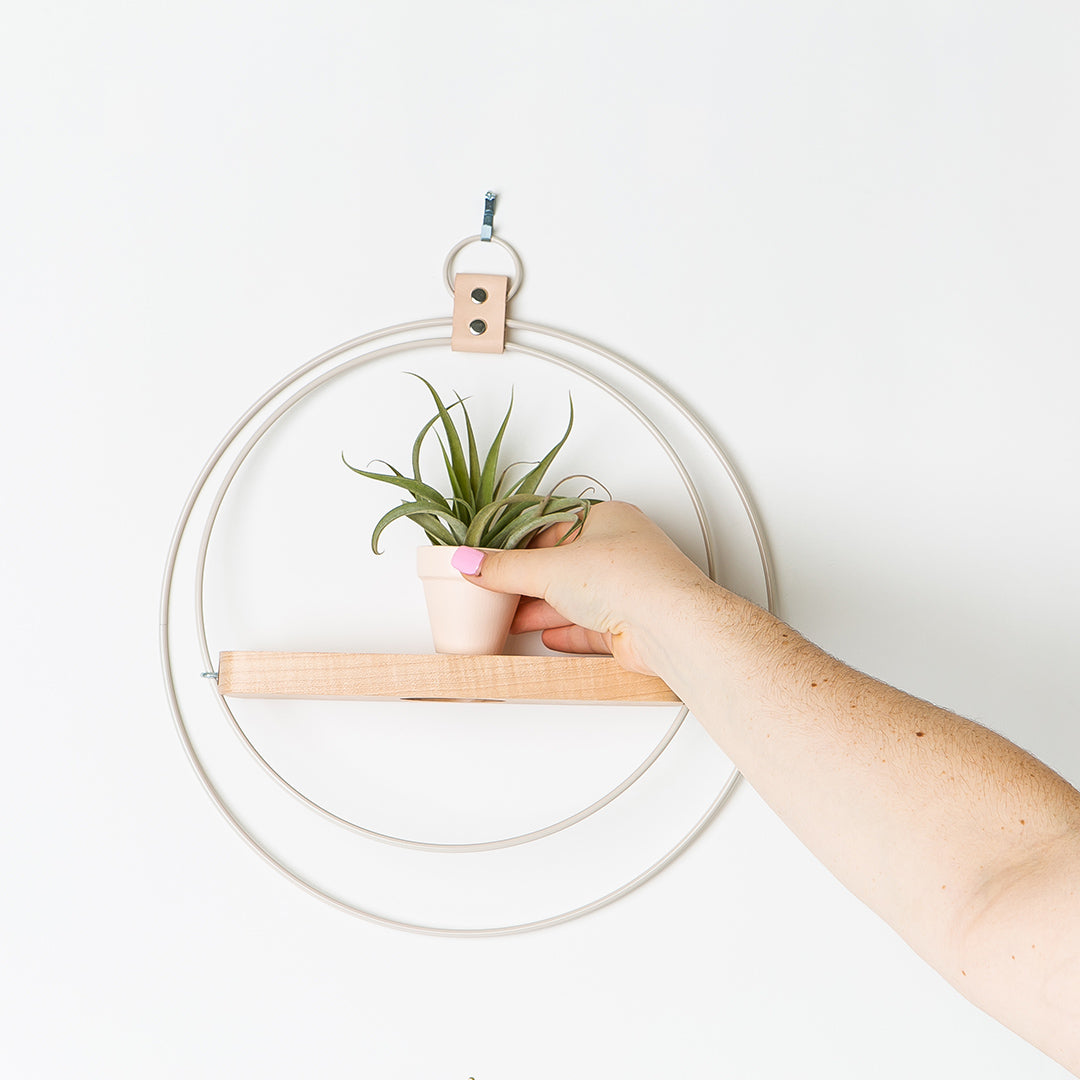 person placing a plant on plant shelf with white metal and maple wood base with blush colored pot